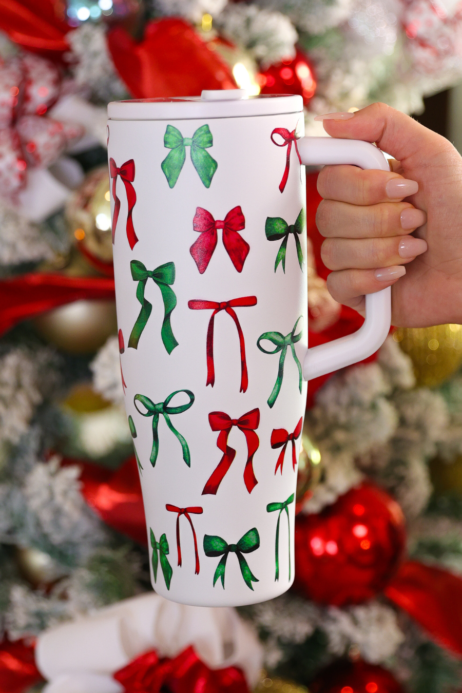 White tumbler with red and green bow pattern held in front of a decorated Christmas tree.