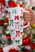White tumbler with red and green bow pattern held in front of a decorated Christmas tree.